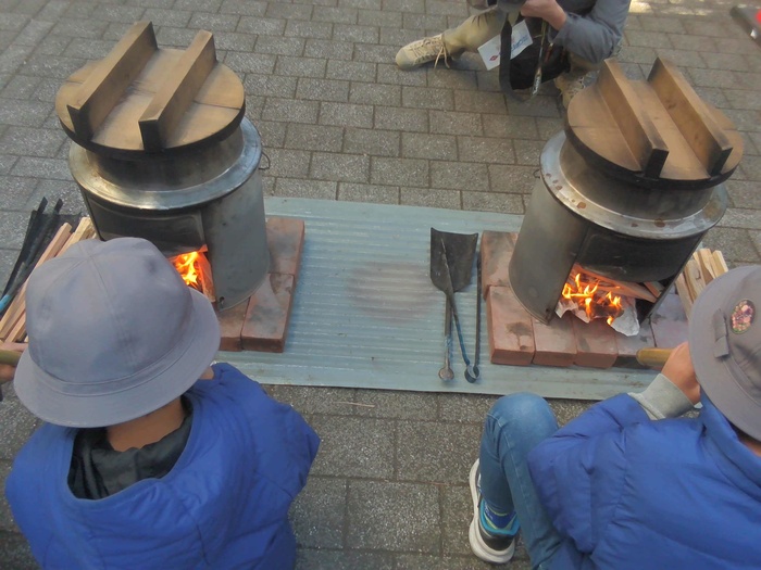 子どもたちがかまど体験でかまどの火に空気を送り込んでいる写真